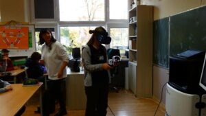 Na zdjęciu widzimy lekcję chemii. Uczennica w goglach VR i kontrolerami na rękach, prowadzi doświadczenie w świecie wirtualnym