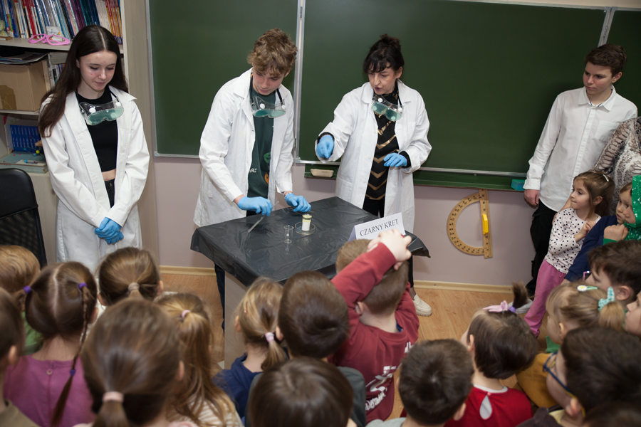 Na zdjęciu, grupa przedszkolaków oraz nauczyciel chemii wraz z 3 uczniów uczniami klas starszych prezentujący doświadczenia chemiczne
